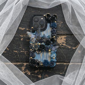 May include: A decorative phone case featuring a blue and black floral design with butterfly accents. The case is adorned with sculpted roses, butterflies, and gold-colored embellishments. The case is designed to fit a smartphone.