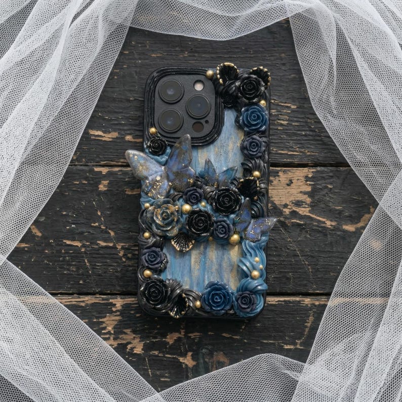 May include: A decorative phone case featuring a blue and black floral design with butterfly accents. The case is adorned with sculpted roses, butterflies, and gold-colored embellishments. The case is designed to fit a smartphone.
