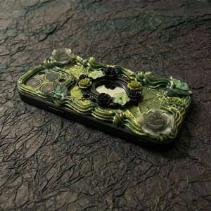 Op de afbeelding: Een zwarte smartphonehoes versierd met een getextureerd, groen, wervelend glazuurachtig ontwerp. De hoes is voorzien van diverse kleine, driedimensionale roosvormige versieringen in zwart, groen en doorschijnend grijs, met een centrale ronde spiegel.