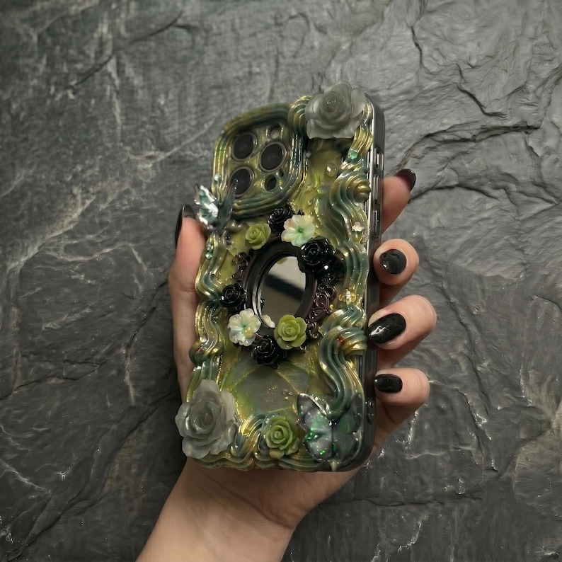 Op de afbeelding: Een decoratief telefoonhoesje met een wervelende geel-groene harsbasis, versierd met driedimensionale bloemaccenten in wit, groen en zwart, twee iriserende vlinderfiguren en een kleine centrale ronde spiegel.