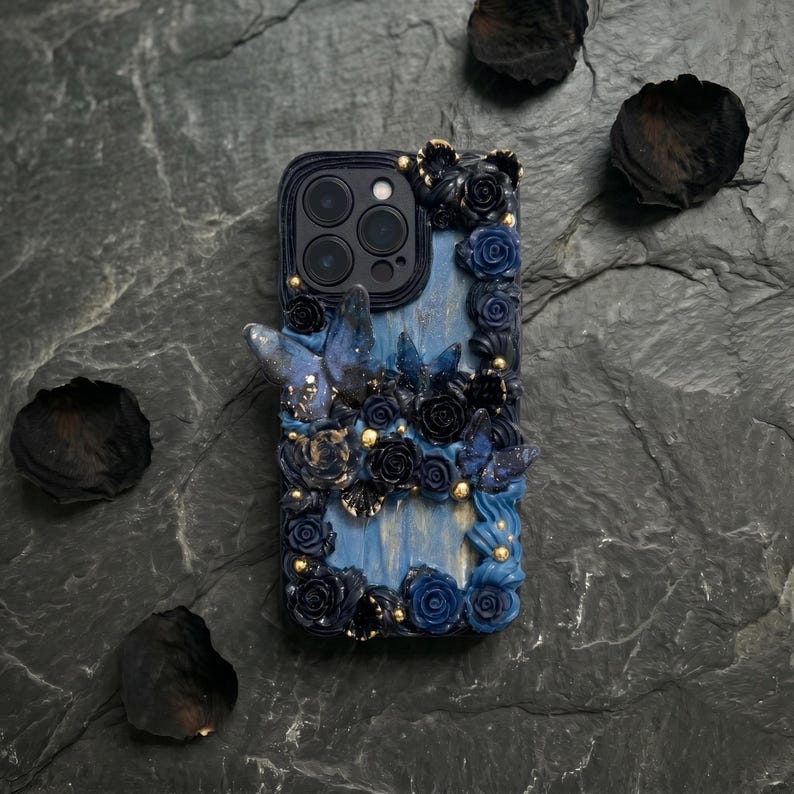 May include: A decorative phone case with a blue and black floral design. The case features sculpted roses, butterflies, and gold accents. The case is designed to fit a smartphone and has a camera cutout.
