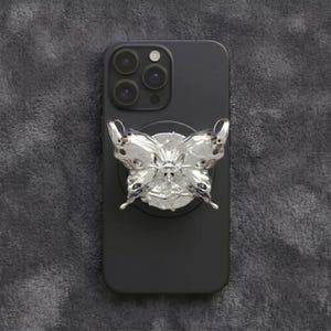 May include: A black smartphone with a metallic silver butterfly-shaped phone grip attached to the centre of the back. The butterfly has a smooth, reflective, liquid-like surface with intricate wing details and is mounted on a circular base.