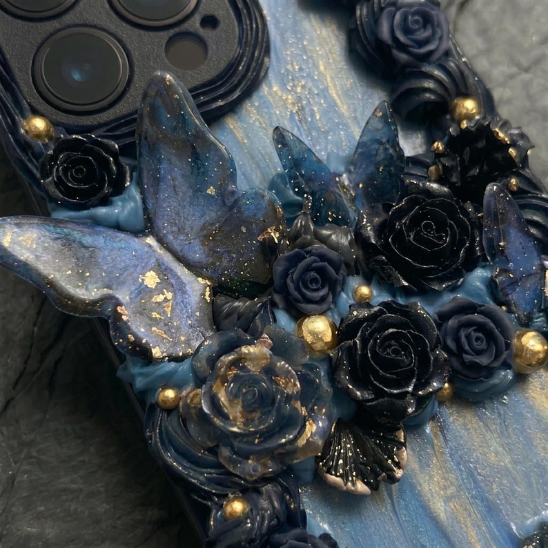 May include: A custom phone case featuring a design of blue butterflies, black roses, and gold accents. The case has a dark blue base with a textured, flowing pattern. The butterflies and roses are intricately detailed, adding a touch of elegance.