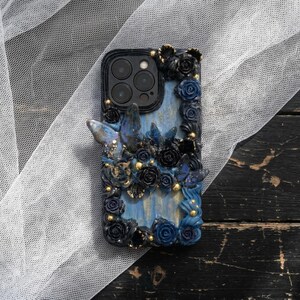 May include: A decorative phone case featuring a blue and black floral design with butterfly accents. The case has a black border and a light blue background. Gold-colored beads are interspersed throughout the floral arrangement.