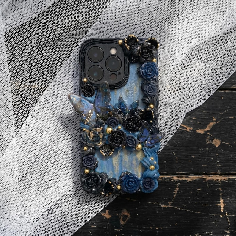 May include: A decorative phone case featuring a blue and black floral design with butterfly accents. The case has a black border and a light blue background. Gold-colored beads are interspersed throughout the floral arrangement.