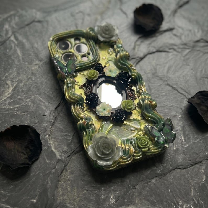 Op de afbeelding: Een decoratief telefoonhoesje met een geel en groen gemarmerde basis en verhoogde, golvende groene accenten. Het ontwerp bevat diverse driedimensionale bloemversieringen in wit, zwart en groen, samen met twee vlinderfiguren.
