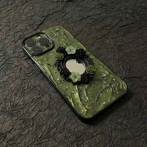 Op de afbeelding: Een telefoonhoesje met een getextureerd olijfgroen oppervlak, voorzien van een centrale ovale spiegel omringd door kleine zwarte en lichtgroene bloemversieringen. Het hoesje is gedecoreerd met verspreide veelkleurige strass-steentjes en heeft een uitsparing voor drie camera's.
