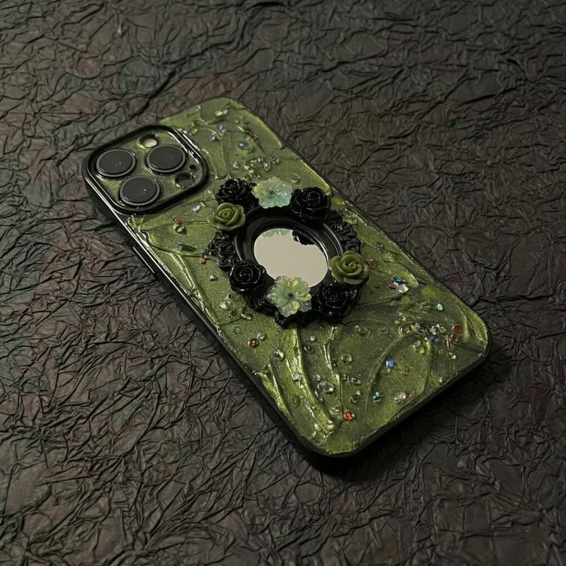 Op de afbeelding: Een telefoonhoesje met een getextureerd olijfgroen oppervlak, voorzien van een centrale ovale spiegel omringd door kleine zwarte en lichtgroene bloemversieringen. Het hoesje is gedecoreerd met verspreide veelkleurige strass-steentjes en heeft een uitsparing voor drie camera's.