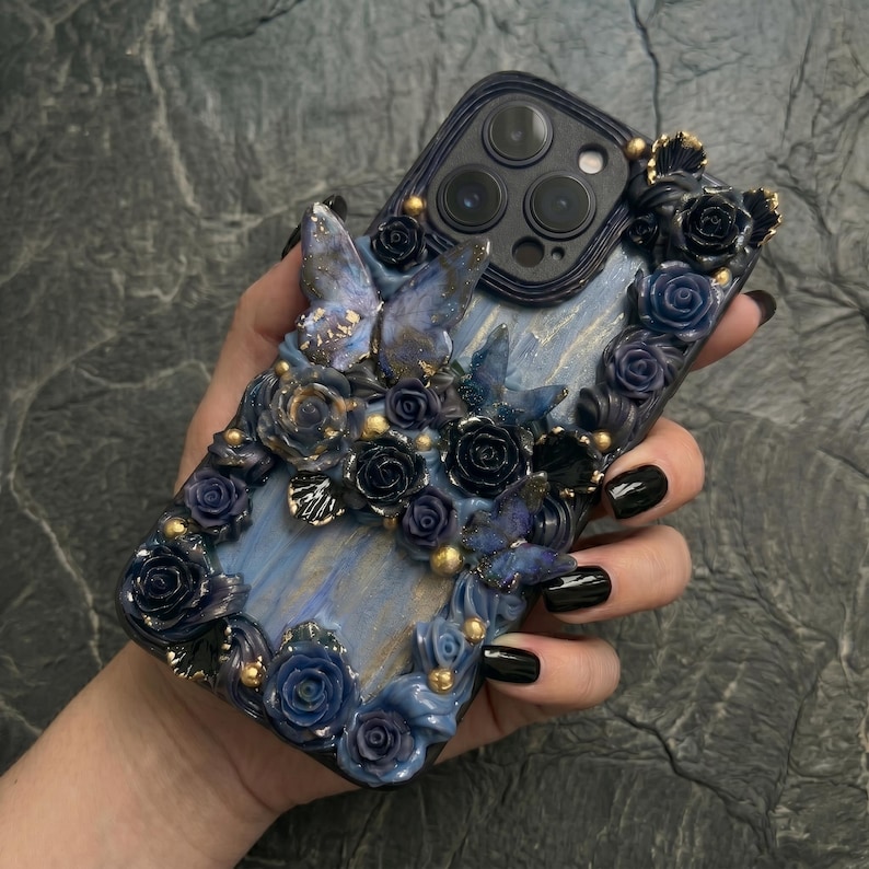 May include: A decorative phone case featuring a blue and gold floral design with butterfly accents. The case is adorned with sculpted roses and gold embellishments, creating a luxurious and artistic aesthetic. The case is designed for a smartphone.