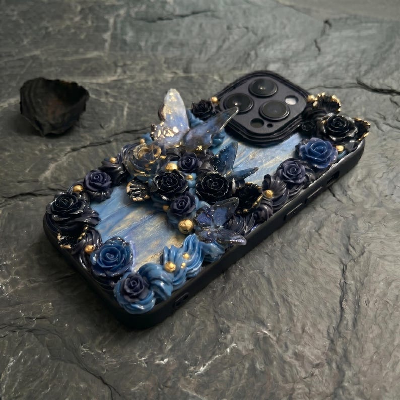 May include: A black phone case with a decorative floral design. The case features blue and black roses, butterflies, and gold accents. The background has a blue marbled effect, creating a unique and artistic look.