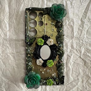 Op de afbeelding: Een rechthoekig telefoonhoesje met een goudkleurige, getextureerde achtergrond en versieringen van donkergroene, zwarte en limoengroene bloemen. Het bevat een centrale ovale spiegel omringd door kleine bloemetjes en twee grote, doorschijnende groene rozen.