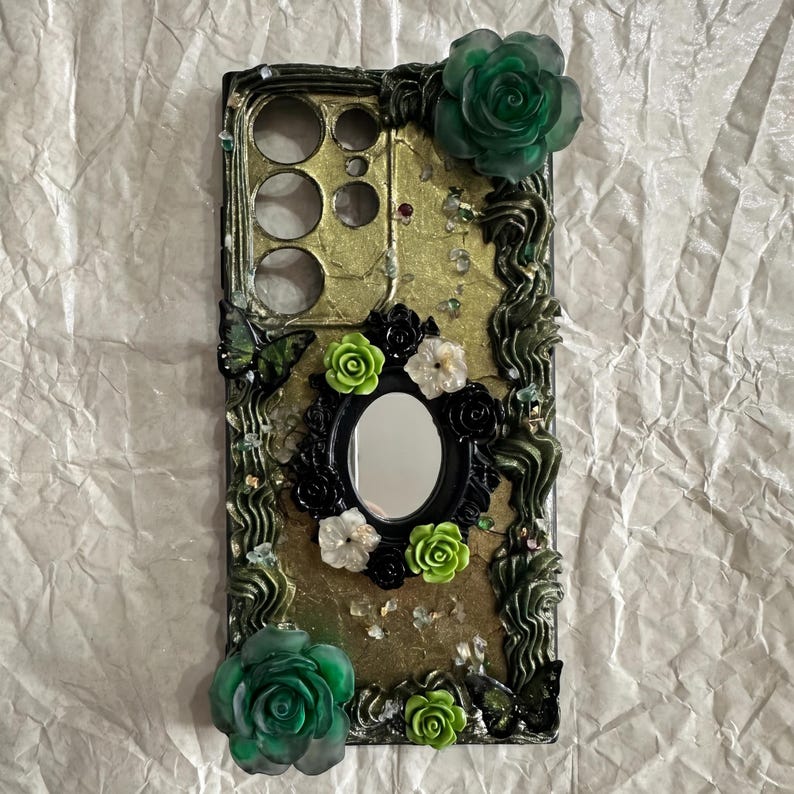 Op de afbeelding: Een rechthoekig telefoonhoesje met een goudkleurige, getextureerde achtergrond en versieringen van donkergroene, zwarte en limoengroene bloemen. Het bevat een centrale ovale spiegel omringd door kleine bloemetjes en twee grote, doorschijnende groene rozen.