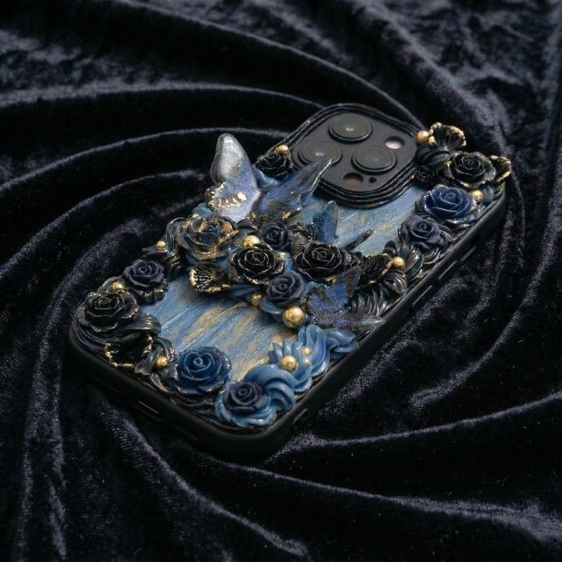 May include: Black phone case with a detailed floral and butterfly design. The case features blue and black roses, butterflies, and gold accents. The design is set against a blue and gold marbled background.