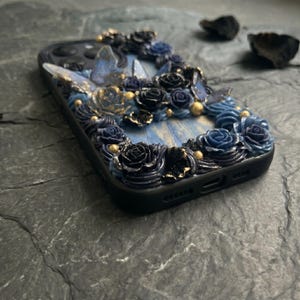 May include: Black phone case decorated with blue and black roses, gold accents, and butterfly details. The case has a textured, floral design with a light blue background.