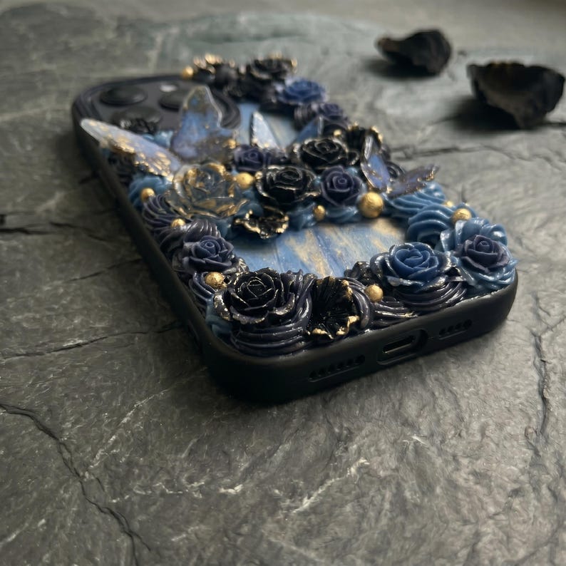 May include: Black phone case decorated with blue and black roses, gold accents, and butterfly details. The case has a textured, floral design with a light blue background.