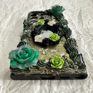 Op de afbeelding: Een zwarte smartphonehoes met een gestructureerde goudkleurige glitterbasis, versierd met driedimensionale groene en zwarte harsrozen, een kleine centrale ronde spiegel en een zwart vlinderaccent. De randen zijn gedecoreerd met donkergroene hars.