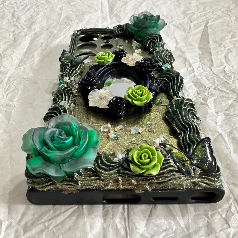 Op de afbeelding: Een zwarte smartphonehoes met een gestructureerde goudkleurige glitterbasis, versierd met driedimensionale groene en zwarte harsrozen, een kleine centrale ronde spiegel en een zwart vlinderaccent. De randen zijn gedecoreerd met donkergroene hars.