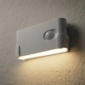 May include: A white, rectangular motion-activated LED light fixture mounted on a textured wall. The fixture has a built-in sensor and a horizontal light strip emitting a warm glow. The design is modern and minimalist.