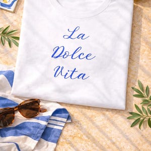 Könnte beinhalten: Weißes T-Shirt mit dem blauen Schriftzug "La Dolce Vita". Das Shirt liegt auf einem gestreiften blau-weißen Handtuch, mit Sonnenbrille, Zitronen und Grünzeug. Die Szene spielt sich auf sandigem Untergrund ab.