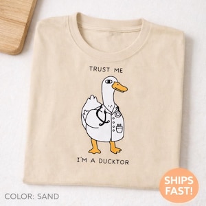 Könnte beinhalten: Sandfarbenes T-Shirt mit einer Cartoon-Ente im Arztkittel, komplett mit Stethoskop. Der Text "TRUST ME" steht über der Ente, und "I'M A DUCKTOR" darunter. Das T-Shirt ist sauber gefaltet.