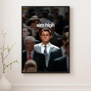 May include: Framed print with the text "aim high" in white, featuring a man in a suit. The background is blurred, with other people. The print is displayed on a white wall, with a vase and plant in the foreground.