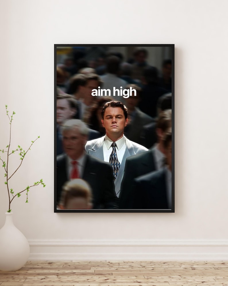 May include: Framed print with the text "aim high" in white, featuring a man in a suit. The background is blurred, with other people. The print is displayed on a white wall, with a vase and plant in the foreground.