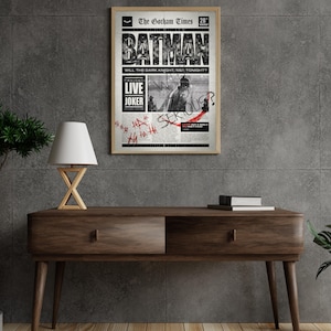 Może przedstawiać: Oprawiony plakat z motywem gazety w stylu vintage, z nagł&oacute;wkiem "BATMAN" pogrubioną czcionką. Plakat zawiera tekst "LIVE JOKER" i grafikę Batmana. Plakat jest wyświetlany na drewnianej konsoli.