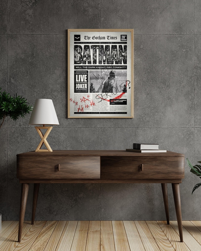 Może przedstawiać: Oprawiony plakat z motywem gazety w stylu vintage, z nagł&oacute;wkiem "BATMAN" pogrubioną czcionką. Plakat zawiera tekst "LIVE JOKER" i grafikę Batmana. Plakat jest wyświetlany na drewnianej konsoli.
