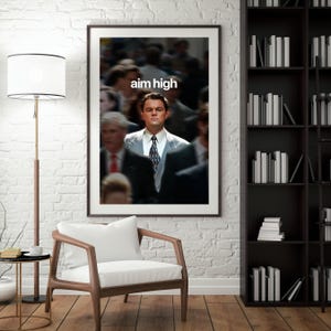 May include: A framed print with the words "aim high" above a man in a suit. The image has a blurred background of people. The artwork is displayed in a room with a white brick wall, a wooden chair, and a bookshelf filled with books.