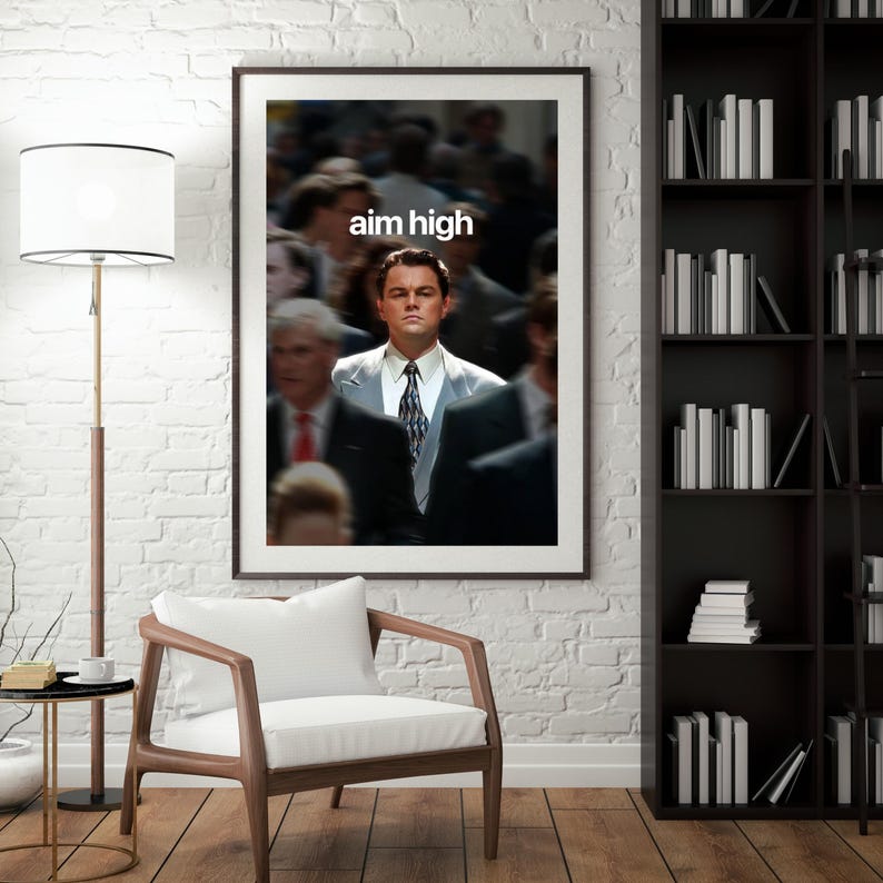 May include: A framed print with the words "aim high" above a man in a suit. The image has a blurred background of people. The artwork is displayed in a room with a white brick wall, a wooden chair, and a bookshelf filled with books.