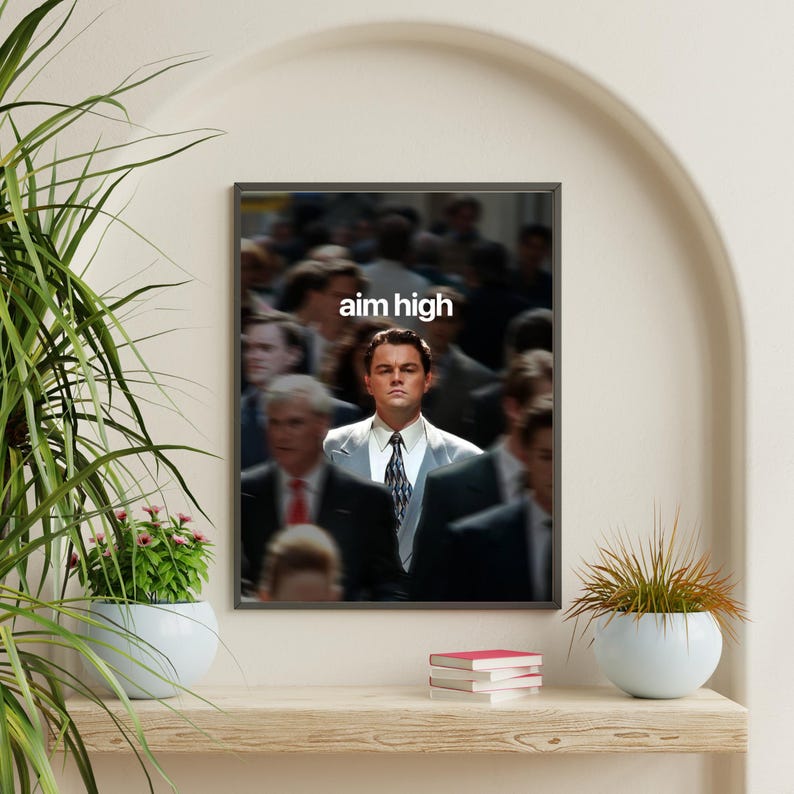 May include: Framed print showing a man in a suit, set against a blurred background of people. The white text "aim high" is above the man's head. The print is displayed on a shelf with plants and books.