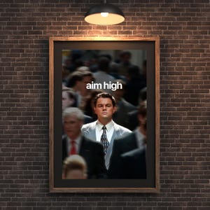 May include: Framed poster with the text "aim high" above a man in a suit, surrounded by blurred figures. The poster is set against a dark brick wall, with a hanging light fixture above.
