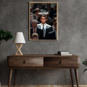 May include: A framed print with a blurred background of people and a man in a suit in focus. The text "aim high" is displayed in white at the top. The print is above a wooden console table with a lamp.