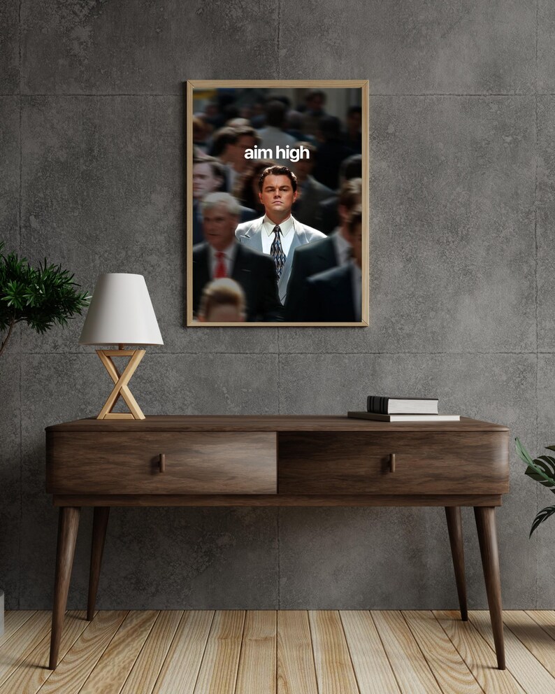 May include: A framed print with a blurred background of people and a man in a suit in focus. The text "aim high" is displayed in white at the top. The print is above a wooden console table with a lamp.