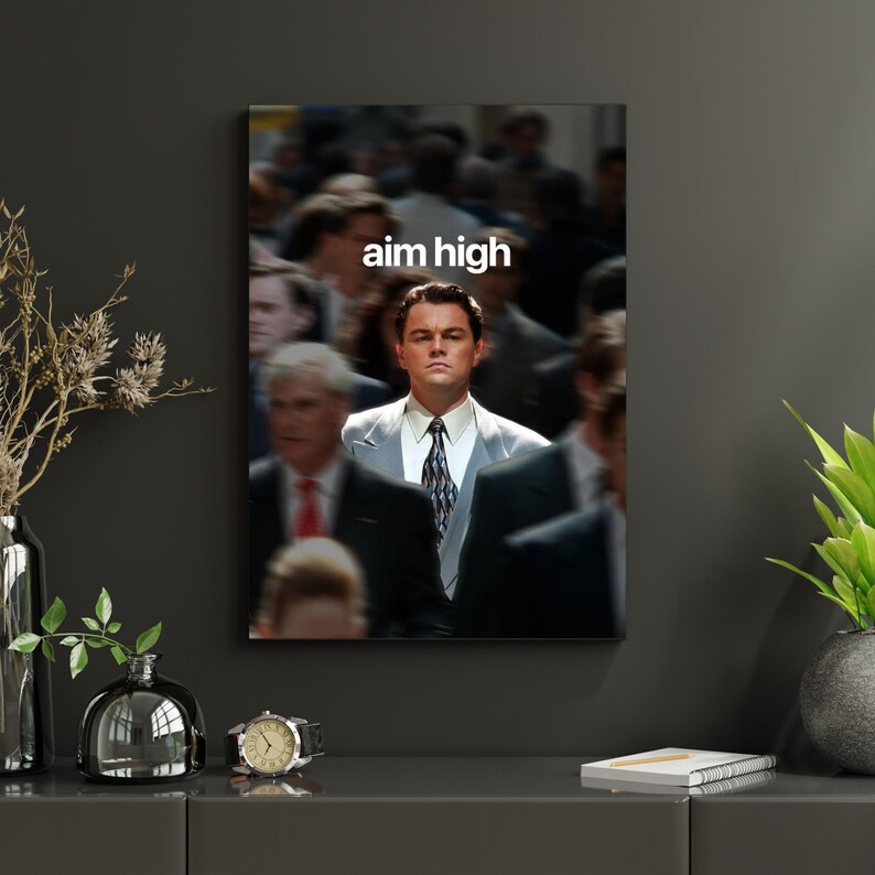 May include: A framed print showing a man in a suit, set against a blurred background of people. The words "aim high" are printed in white. The image has a cinematic quality, with the central figure in sharp focus.