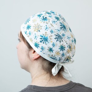 May include: A white scrub cap featuring a pattern of blue and gold snowflakes. The cap is tied at the back with a white ribbon. The cap is worn by a person.