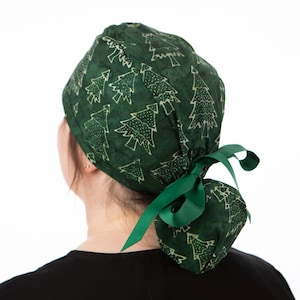 May include: A dark green scrub cap featuring a white Christmas tree design. The cap is tied at the back with a green ribbon. The person is wearing a black top.