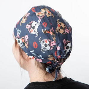 May include: A blue surgical scrub hat with a repeating pattern of cartoon dogs with pink lips and tongues sticking out. The text "Kiss Me" is printed on the hat.