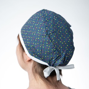Op de afbeelding: Een marineblauwe operatiemuts met witte rand en striksluiting. De muts is bedrukt met kleine, kleurrijke confetti-achtige vormen in rood, geel en groen. Ontworpen voor gebruik in een medische omgeving.