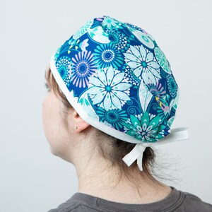 May include: A blue and white floral patterned scrub cap with a white trim and ties. The cap features a repeating pattern of stylized flowers in shades of blue, teal, and white. The cap is designed to be worn in a medical or healthcare setting.
