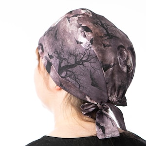 May include: A dark purple and gray Halloween-themed scrub cap. The cap features a print of bats, bare trees, and a full moon. The cap is tied in a bow at the back.