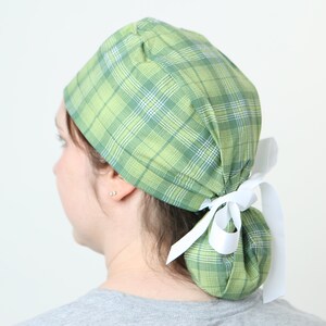 May include: A green plaid scrub cap with a white ribbon tie. The cap is made of a soft fabric and features a classic plaid pattern in shades of green, white, and blue. The cap is tied in a bun at the back.