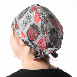 May include: A gray scrub hat with a red and black anatomical heart print. The hat is tied at the back of the head.