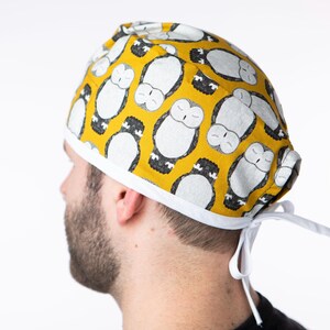 Op de afbeelding: Een gele scrub-muts met een witte rand en een herhalend patroon van slapende uilen. De muts heeft een wit lint dat aan de achterkant van de nek kan worden vastgebonden.