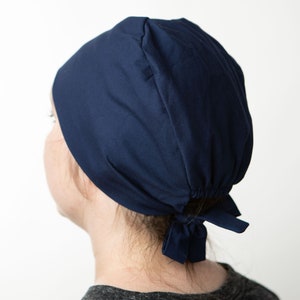 Puede incluir: Un gorro quirúrgico azul marino con la espalda fruncida y cierre de lazo. El gorro está hecho de una tela suave y está diseñado para cubrir el cabello. El fondo es blanco liso.