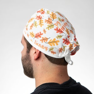 May include: White scrub hat with a pattern of orange and brown leaves. The hat has the text "Happy" and "Giving" printed on it.