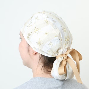 May include: A white and gold patterned scrub cap with a gold ribbon tie. The cap features a repeating pattern of gold snowflakes and is tied at the back. The cap is worn by a person.