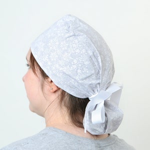 May include: A light grey surgical scrub cap featuring a white floral pattern. The cap is secured at the back with a white ribbon. Designed to cover hair during medical procedures.