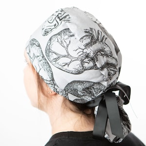 May include: A light gray surgical scrub cap featuring a black anatomical heart and brain print. The cap is tied in the back with a black ribbon. The medical-themed headwear is designed for healthcare professionals.