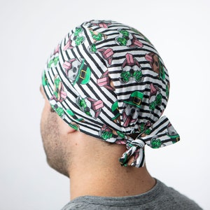 May include: A black and white striped scrub hat with a green shamrock and cartoon dog design. The hat has a tie at the back.